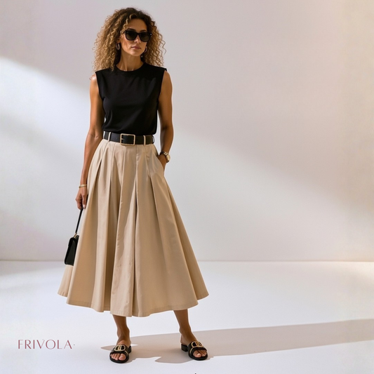 A woman models a stylish outfit featuring a sleeveless black top and a high-waisted, pleated beige midi skirt with a black belt what is actually a one piece dress. She is wearing black slide sandals and holding a small black handbag, set against a minimalist studio background with soft shadows.